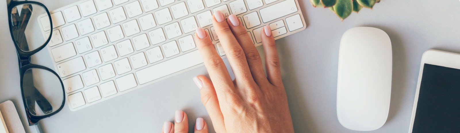 a hand on a keyboard, reading glasses on the left, a computer mouse and mobile phone on the right